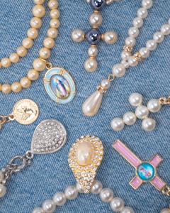 A collection of religious-themed jewelry arranged on a blue denim fabric. The pieces include beaded rosaries, pendants, and medals featuring pearls and gold accents, with some displaying religious icons and symbols.