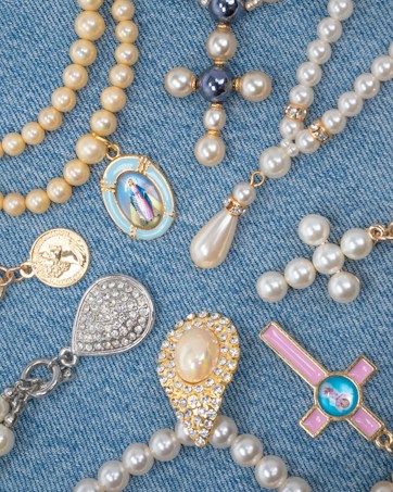 A collection of religious-themed jewelry arranged on a blue denim fabric. The pieces include beaded rosaries, pendants, and medals featuring pearls and gold accents, with some displaying religious icons and symbols.