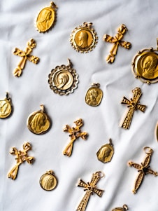 A collection of gold religious medallions and crosses are arranged on a white fabric background. The medallions feature various religious figures and intricate designs, while the crosses have detailed engravings. The layout suggests a sense of careful arrangement, highlighting the craftsmanship of each piece.