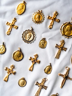 A collection of gold religious medallions and crosses are arranged on a white fabric background. The medallions feature various religious figures and intricate designs, while the crosses have detailed engravings. The layout suggests a sense of careful arrangement, highlighting the craftsmanship of each piece.