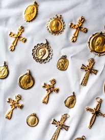 A collection of gold religious medallions and crosses are arranged on a white fabric background. The medallions feature various religious figures and intricate designs, while the crosses have detailed engravings. The layout suggests a sense of careful arrangement, highlighting the craftsmanship of each piece.