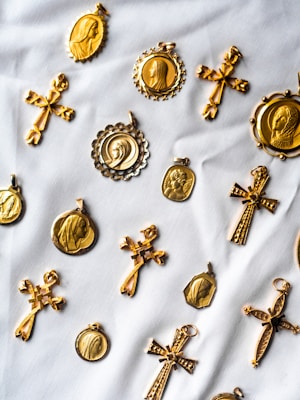 A collection of gold religious medallions and crosses are arranged on a white fabric background. The medallions feature various religious figures and intricate designs, while the crosses have detailed engravings. The layout suggests a sense of careful arrangement, highlighting the craftsmanship of each piece.