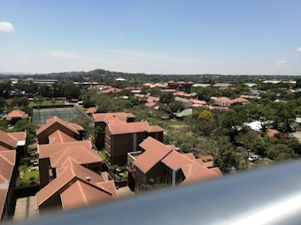 A suburban neighborhood with numerous houses featuring reddish-brown roofs. The view extends to a tennis court surrounded by trees. The landscape is lush with greenery and extends towards a distant hill under a clear blue sky.