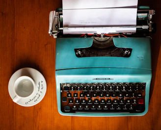 green and black typewriter beside white ceramic mug on brown wooden table