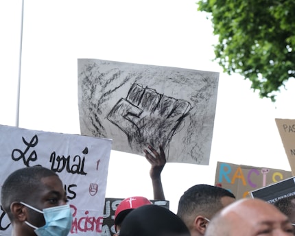 A group of people holds up protest signs. One prominent sign features a charcoal or pencil drawing of a raised fist, symbolizing solidarity and resistance, with words like 'Justice' and 'Fier' written on it. Other signs contain text in different languages, pointing to themes of equality and anti-racism. The crowd includes individuals wearing masks, and the scene is set outdoors with a leafy tree visible in the background.