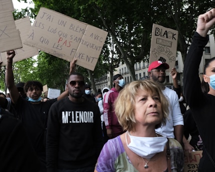 A group of people participating in a protest, holding signs with messages. Some wear masks, and the signs carry statements and slogans. The setting appears to be outdoors with trees and buildings in the background.