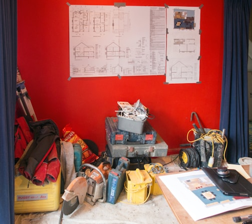 A room with a vivid red wall featuring various blueprints and architectural drawings mounted on it. The floor is cluttered with construction tools and supplies, including boxes, cables, and a saw. A table nearby holds additional papers and a lens cap.