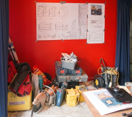 A room with a vivid red wall featuring various blueprints and architectural drawings mounted on it. The floor is cluttered with construction tools and supplies, including boxes, cables, and a saw. A table nearby holds additional papers and a lens cap.