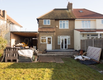 A semi-detached house with a brown pebble-dash exterior and white-framed windows stands in the middle of the image. The backyard appears to be under construction or renovation, with building materials such as bricks, wood pallets, and tarps scattered across the lawn. A garage or carport area to the left is partially covered and houses some construction equipment. A wooden fence borders the right side of the property, and the grass shows signs of recent activity.