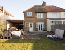 A semi-detached house with a brown pebble-dash exterior and white-framed windows stands in the middle of the image. The backyard appears to be under construction or renovation, with building materials such as bricks, wood pallets, and tarps scattered across the lawn. A garage or carport area to the left is partially covered and houses some construction equipment. A wooden fence borders the right side of the property, and the grass shows signs of recent activity.