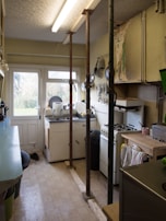A narrow kitchen features a sink and countertop near a window with light streaming in. A stove is visible to the right, and several metal poles are installed vertically from floor to ceiling. The walls show signs of damage with peeling paint and cracks. Utensils and kitchen tools hang from a wall rack, and various kitchen items are scattered on the countertops.