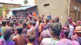 A large group of people are gathered in a vibrant celebration. Bright colors cover their clothes, skin, and hair, suggesting a festival involving powder or paint. The setting appears to be a narrow street in a residential area, with buildings visible in the background. Everyone seems to be engaged and enjoying the festive activity.