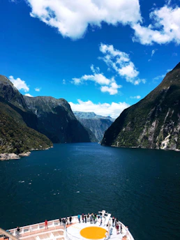 A cruise ship deck with several people standing and observing the surrounding scenery. The ship is navigating through a narrow waterway, flanked by towering, lush green mountains under a vibrant blue sky with scattered clouds.