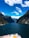 A cruise ship deck with several people standing and observing the surrounding scenery. The ship is navigating through a narrow waterway, flanked by towering, lush green mountains under a vibrant blue sky with scattered clouds.