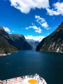 A cruise ship deck with several people standing and observing the surrounding scenery. The ship is navigating through a narrow waterway, flanked by towering, lush green mountains under a vibrant blue sky with scattered clouds.