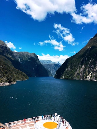 A cruise ship deck with several people standing and observing the surrounding scenery. The ship is navigating through a narrow waterway, flanked by towering, lush green mountains under a vibrant blue sky with scattered clouds.