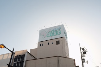 A large building topped with a billboard displaying the word 'Value' in stylized blue and white text. The sky is clear and slightly illuminated by the setting or rising sun. A streetlamp is visible on the left, with utility poles and wires on the right.