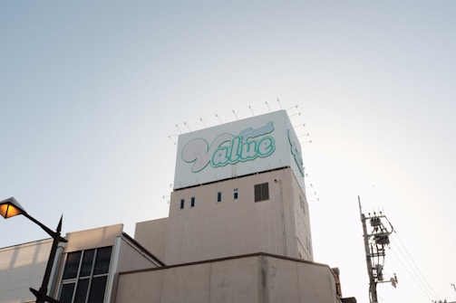 A large building topped with a billboard displaying the word 'Value' in stylized blue and white text. The sky is clear and slightly illuminated by the setting or rising sun. A streetlamp is visible on the left, with utility poles and wires on the right.