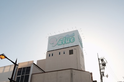 A large building topped with a billboard displaying the word 'Value' in stylized blue and white text. The sky is clear and slightly illuminated by the setting or rising sun. A streetlamp is visible on the left, with utility poles and wires on the right.
