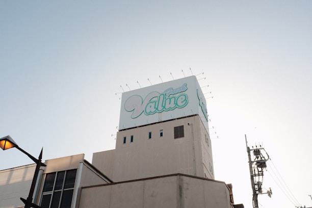 A large building topped with a billboard displaying the word 'Value' in stylized blue and white text. The sky is clear and slightly illuminated by the setting or rising sun. A streetlamp is visible on the left, with utility poles and wires on the right.