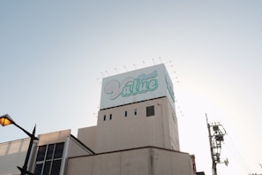 A large building topped with a billboard displaying the word 'Value' in stylized blue and white text. The sky is clear and slightly illuminated by the setting or rising sun. A streetlamp is visible on the left, with utility poles and wires on the right.