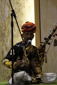 A man dressed in traditional clothing is playing a string instrument while sitting cross-legged. He wears a colorful turban and is positioned in front of a microphone stand, suggesting a performance setting. The textured background with warm lighting enhances the cultural ambiance.