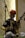 A man dressed in traditional clothing is playing a string instrument while sitting cross-legged. He wears a colorful turban and is positioned in front of a microphone stand, suggesting a performance setting. The textured background with warm lighting enhances the cultural ambiance.