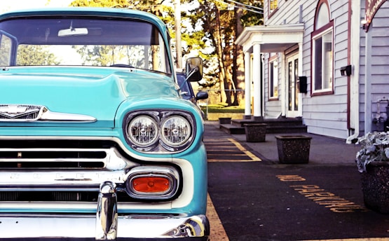 A turquoise vintage car is parked beside a building with white siding and red trim. The foreground focuses on the car's front end, showcasing its distinct headlights and chrome detailing. Flower pots and a 'no parking' sign are visible on the pavement.