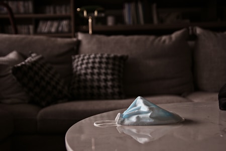 A face mask is placed on a white table in front of a gray couch. The couch has several pillows with a houndstooth pattern. The background suggests a comfortable interior setting with bookshelves and muted lighting.