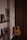 A cozy interior corner featuring a classical guitar leaning against a wooden wall beside a minimalistic wooden shelf. The shelf holds framed photographs, a small potted plant, and a decorative item. Soft lighting casts gentle shadows, creating a warm and intimate atmosphere.