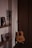 A cozy interior corner featuring a classical guitar leaning against a wooden wall beside a minimalistic wooden shelf. The shelf holds framed photographs, a small potted plant, and a decorative item. Soft lighting casts gentle shadows, creating a warm and intimate atmosphere.