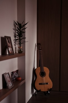 A cozy interior corner featuring a classical guitar leaning against a wooden wall beside a minimalistic wooden shelf. The shelf holds framed photographs, a small potted plant, and a decorative item. Soft lighting casts gentle shadows, creating a warm and intimate atmosphere.