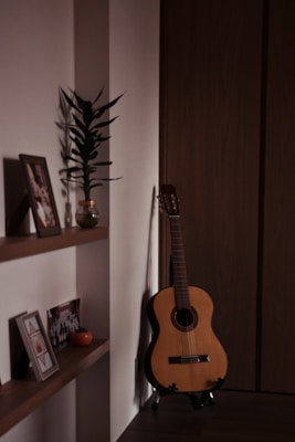 A cozy interior corner featuring a classical guitar leaning against a wooden wall beside a minimalistic wooden shelf. The shelf holds framed photographs, a small potted plant, and a decorative item. Soft lighting casts gentle shadows, creating a warm and intimate atmosphere.