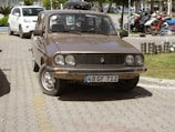 A vintage brown car is parked on a paved road beside a neatly maintained grass lawn. Behind the car, there are several motorcycles parked in an organized manner. A few stacked metal containers are visible on the right side of the image. The car has a rectangular front grille and circular headlights, and a license plate with the markings 48 GF 712 is attached to the front.