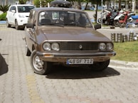 A vintage brown car is parked on a paved road beside a neatly maintained grass lawn. Behind the car, there are several motorcycles parked in an organized manner. A few stacked metal containers are visible on the right side of the image. The car has a rectangular front grille and circular headlights, and a license plate with the markings 48 GF 712 is attached to the front.