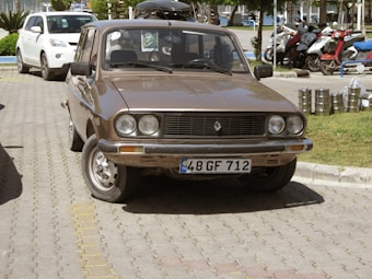 A vintage brown car is parked on a paved road beside a neatly maintained grass lawn. Behind the car, there are several motorcycles parked in an organized manner. A few stacked metal containers are visible on the right side of the image. The car has a rectangular front grille and circular headlights, and a license plate with the markings 48 GF 712 is attached to the front.
