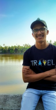 A person wearing a black t-shirt with the word 'TRAVEL' printed on it stands in front of a calm body of water, surrounded by lush green trees. The person is smiling and wearing glasses and a cap.
