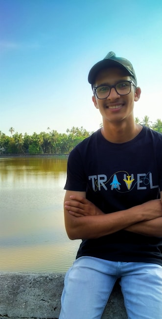 A person wearing a black t-shirt with the word 'TRAVEL' printed on it stands in front of a calm body of water, surrounded by lush green trees. The person is smiling and wearing glasses and a cap.