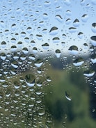 Numerous water droplets are clinging to a clear surface, acting as a lens to the blurred green and blue scenery beyond. Each droplet magnifies and distorts the view, creating a pattern that suggests freshness and natural beauty.