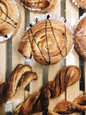 Freshly baked pastries are arranged on a wooden tray, featuring swirls with chocolate and icing drizzles on top. The pastries are golden brown with a flaky texture, and some are twisted in shape, looking crisp and inviting.