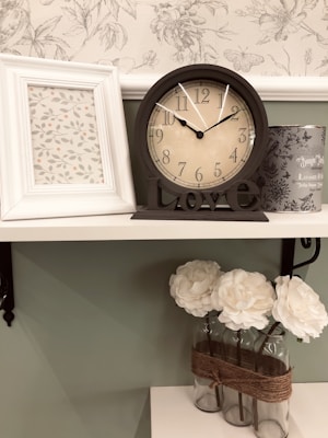 A shelf displaying a decorative arrangement with a framed floral print, an analog clock with the word 'Love' on its base, a cylindrical tin, and a glass vase holding three white flowers. The wallpaper in the background has a subtle, vintage floral pattern.