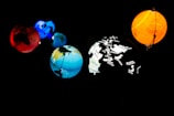 Five planet-like spheres are illuminated against a dark background. The spheres are in various colors: red, blue, orange, and multi-colored with map outlines and corporate names. The arrangement resembles a miniature solar system with each sphere glowing vibrantly.
