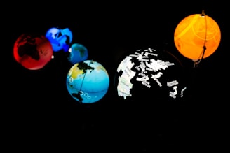Five planet-like spheres are illuminated against a dark background. The spheres are in various colors: red, blue, orange, and multi-colored with map outlines and corporate names. The arrangement resembles a miniature solar system with each sphere glowing vibrantly.