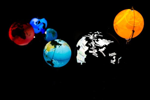 Five planet-like spheres are illuminated against a dark background. The spheres are in various colors: red, blue, orange, and multi-colored with map outlines and corporate names. The arrangement resembles a miniature solar system with each sphere glowing vibrantly.