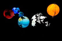 Five planet-like spheres are illuminated against a dark background. The spheres are in various colors: red, blue, orange, and multi-colored with map outlines and corporate names. The arrangement resembles a miniature solar system with each sphere glowing vibrantly.