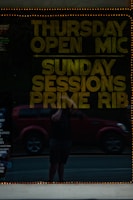 A reflective glass surface displays text promoting events such as Thursday Open Mic and Sunday Sessions Prime Rib. Behind the reflection is a person holding a phone in front of a red vehicle, with the street visible in the background. The glass is bordered by a string of small lights, adding a warm glow.