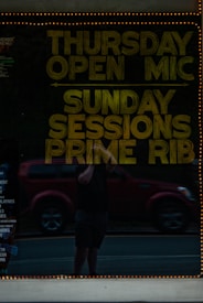 A reflective glass surface displays text promoting events such as Thursday Open Mic and Sunday Sessions Prime Rib. Behind the reflection is a person holding a phone in front of a red vehicle, with the street visible in the background. The glass is bordered by a string of small lights, adding a warm glow.