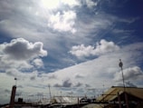 Billowing white clouds spread across a bright blue sky with the sun partially obscured. Various antennas and satellite dishes are mounted on rooftops below, silhouetted against the skyline.