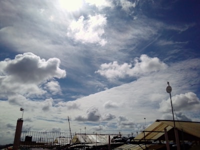 Billowing white clouds spread across a bright blue sky with the sun partially obscured. Various antennas and satellite dishes are mounted on rooftops below, silhouetted against the skyline.