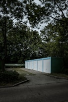A row of five white garage doors with blue frames aligned in a line, set in a wooded environment. The garages are situated on a paved area surrounded by dense greenery, including tall trees with rich foliage. The sky above is overcast, casting a moody ambiance across the scene.